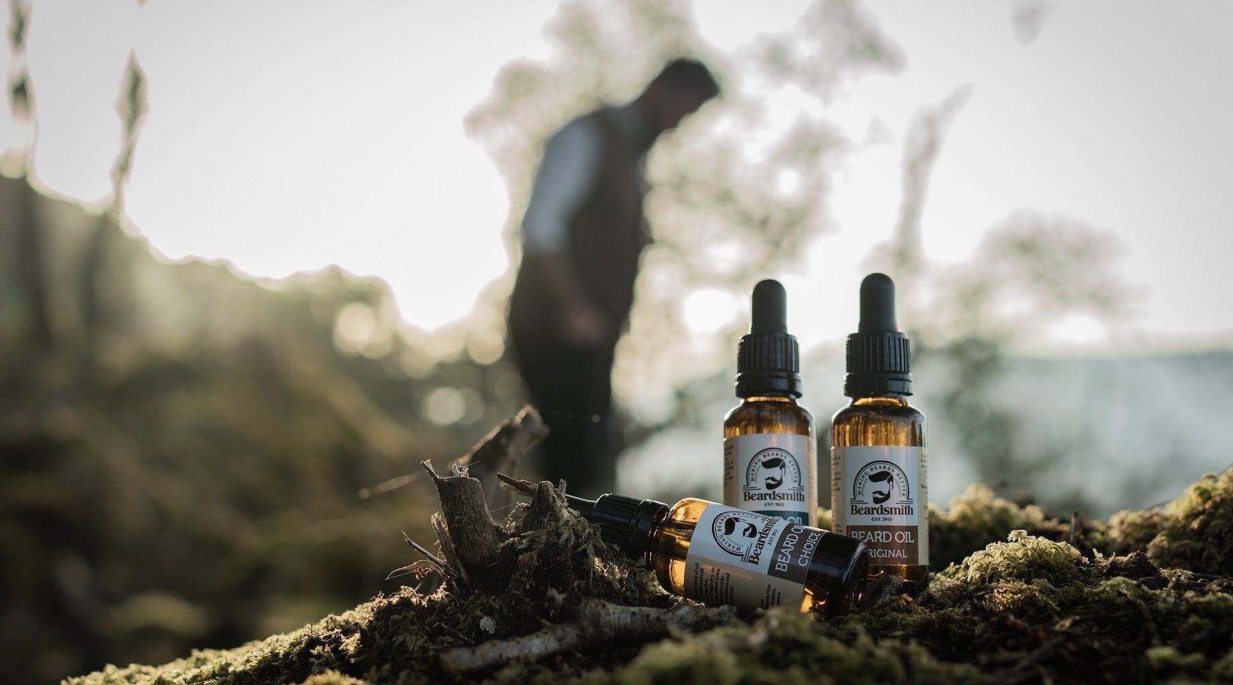 3 bottles of beard oil on a log in a forest with a bearded guys silhouette in the background